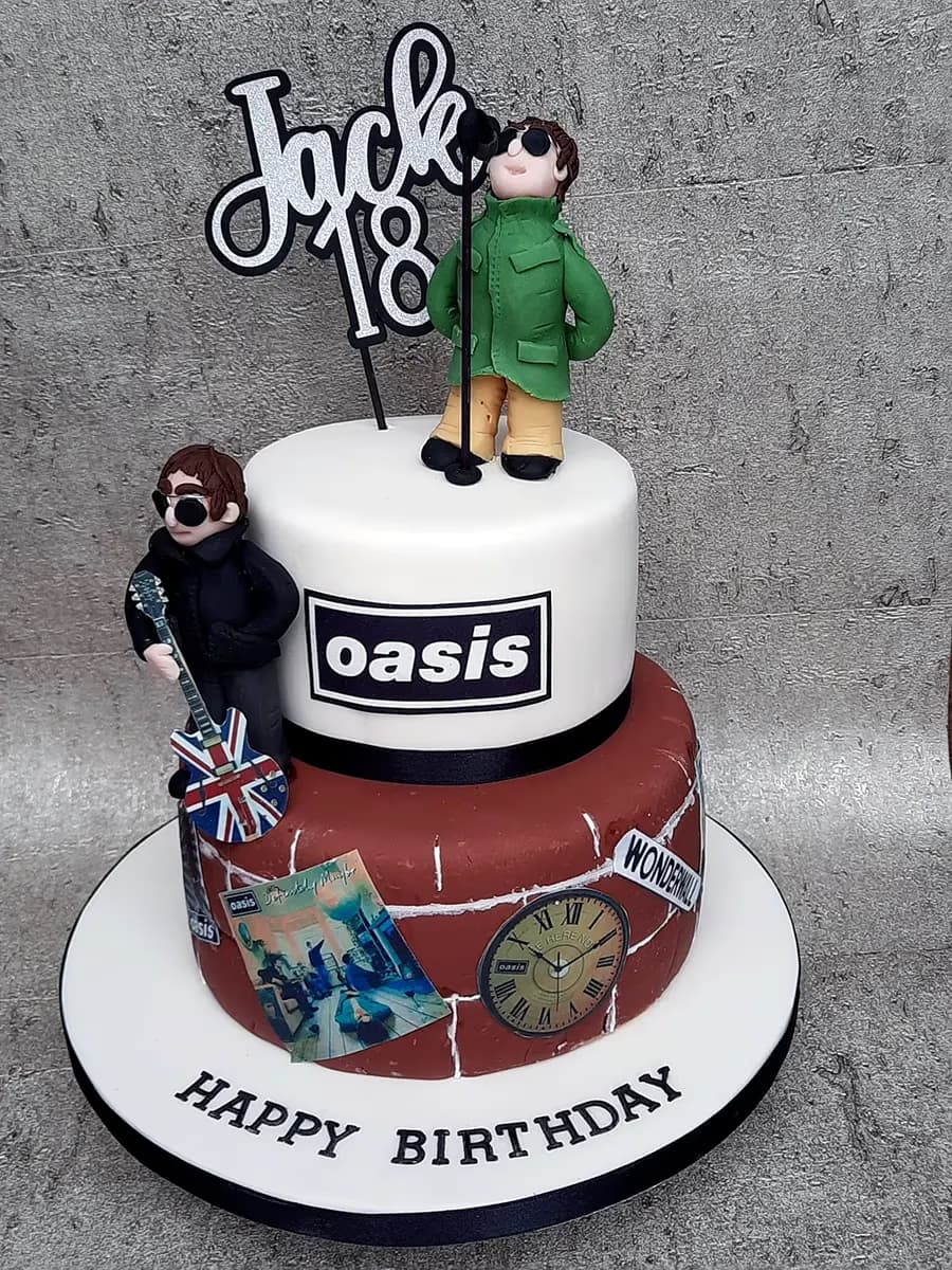 A two-tier Oasis band themed birthday cake for Jack's 18th birthday. The white top tier features an Oasis logo and a fondant figure singing into a microphone, while the reddish-brown bottom tier, designed like a brick wall, has another fondant figure holding a Union Jack guitar, album art, a clock, and a 'Wonderwall' sign. A 'Jack 18' topper is on the cake, and 'HAPPY BIRTHDAY' is written on the white cake board.