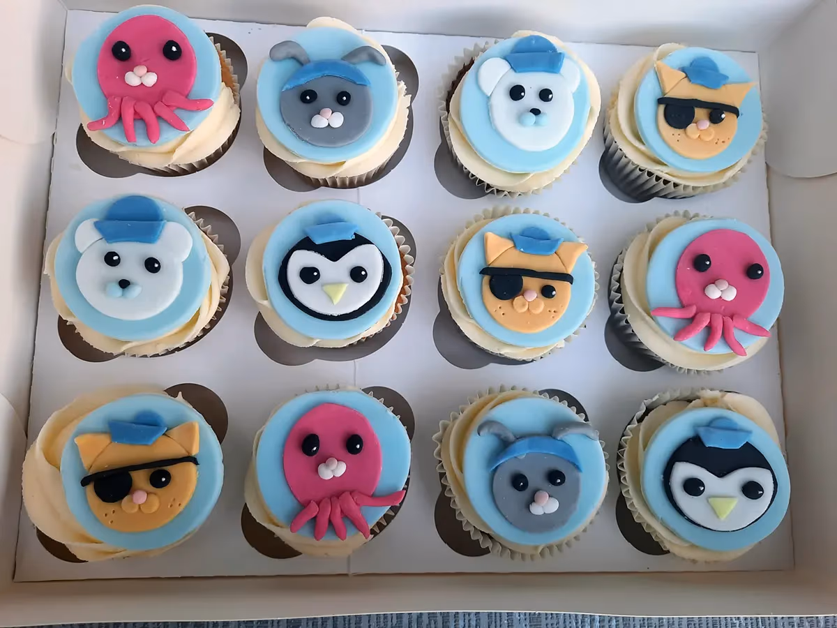 A dozen cupcakes in a white box, each decorated with light blue fondant discs featuring various Octonauts characters including a pink octopus, a white polar bear, a grey rabbit, a black and white penguin, and an orange cat with an eyepatch.