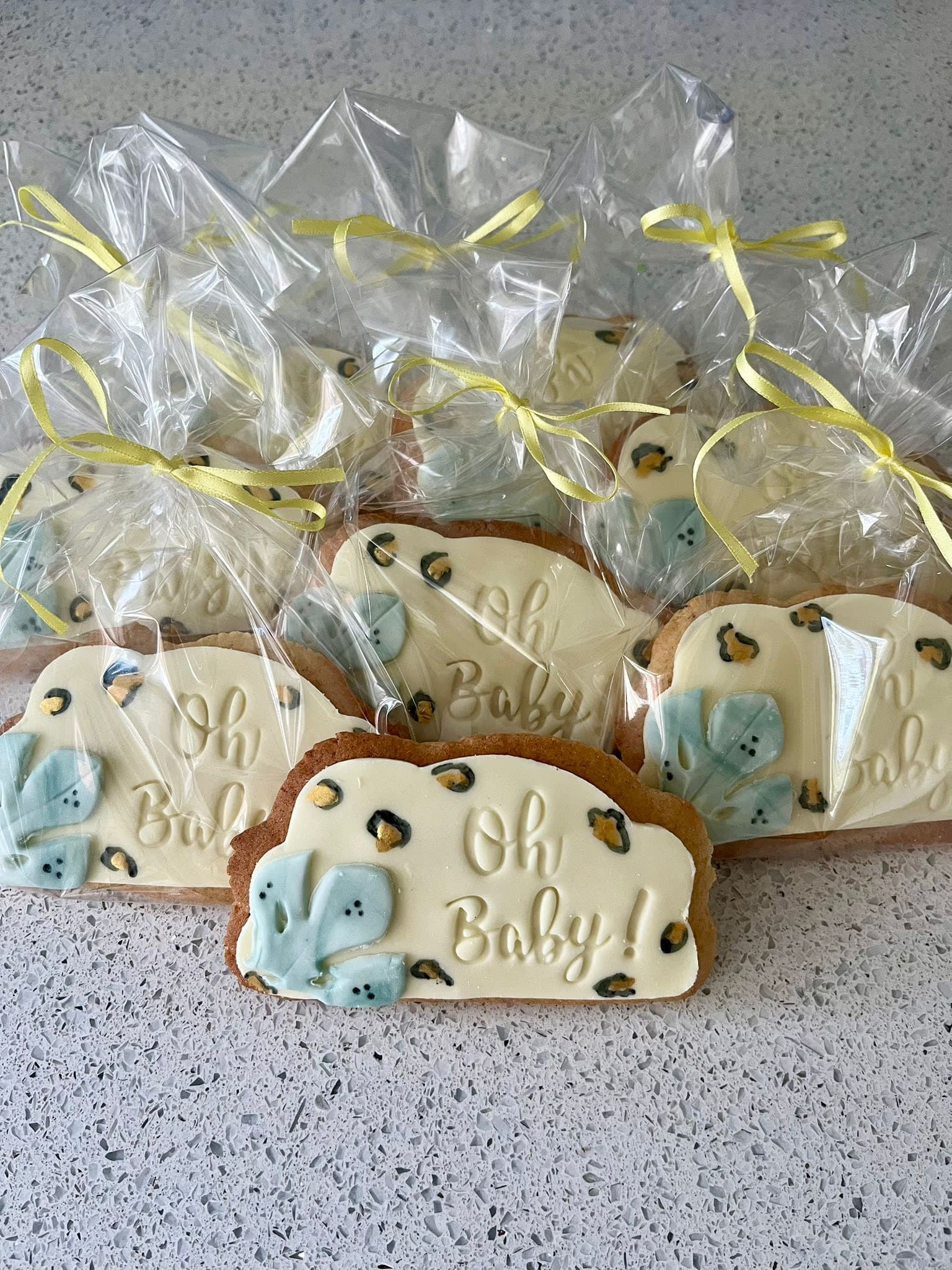 A collection of individually wrapped rectangular cookies in clear plastic bags tied with yellow ribbons. The cookies feature white icing with 'Oh Baby!' text, light blue leaves, and gold and black leopard print spots, arranged on a light grey speckled countertop.