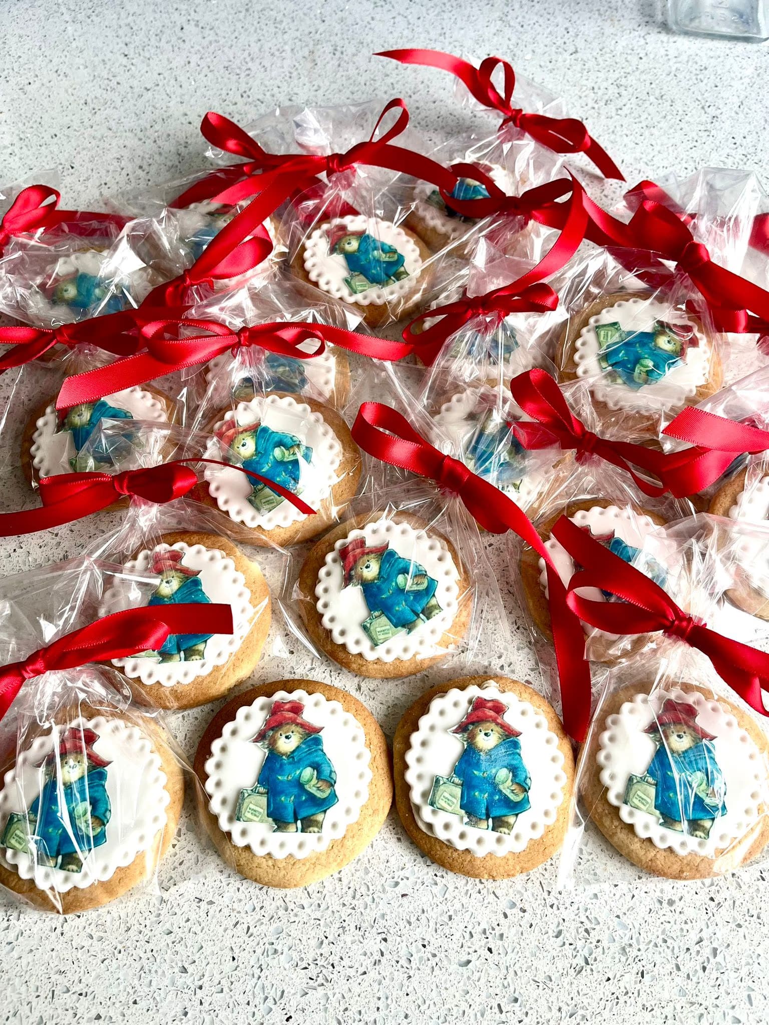 A collection of round cookies, each topped with a white scalloped icing circle featuring an edible image of Paddington Bear, individually wrapped in clear plastic bags tied with red satin ribbons.