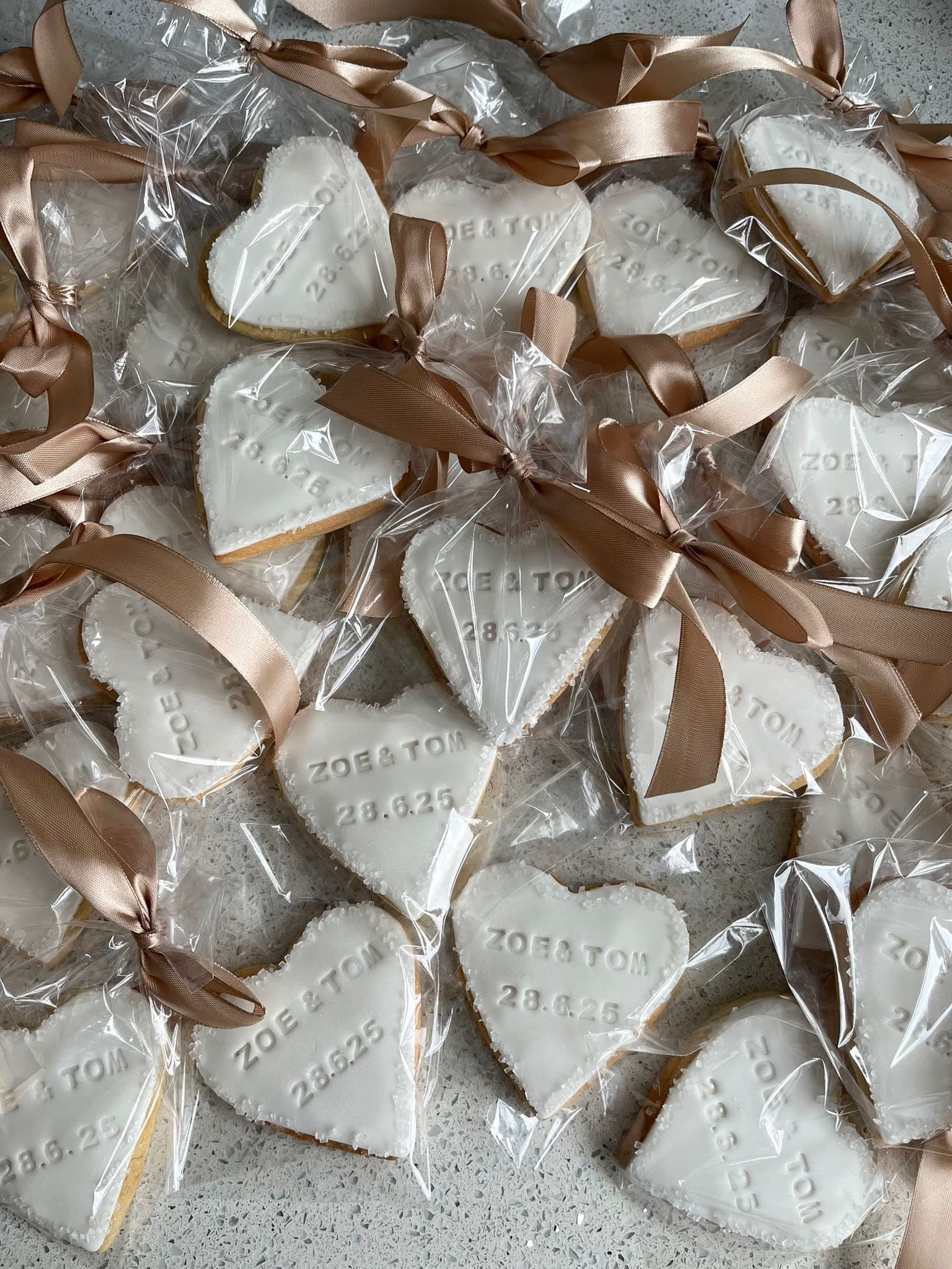 A close-up shot of numerous heart-shaped white iced cookies, individually wrapped in clear plastic and tied with gold ribbons, featuring embossed text 'Zoe & Tom' and '28.6.25'.