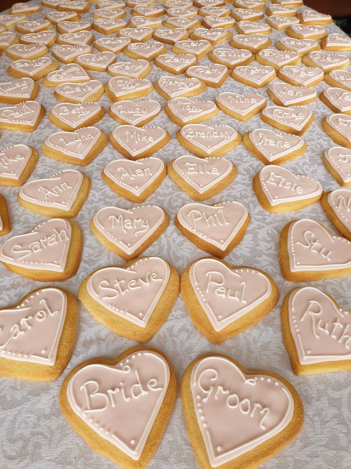 A large collection of heart-shaped cookies with light pink icing and white piped borders, each featuring a different guest name, 'Bride', or 'Groom' written in white script, arranged on a white textured tablecloth.