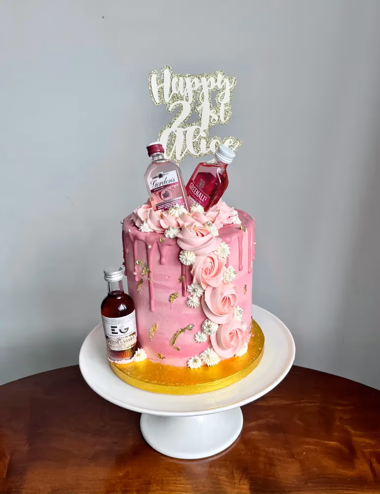 A vibrant pink 21st birthday cake with drip icing, edible gold flakes, pink and white buttercream flowers, and three miniature alcohol bottles (Gordon's, Greenall's, and EG Plum & Vanilla), topped with a glittery 'Happy 21st Alice' cake topper.