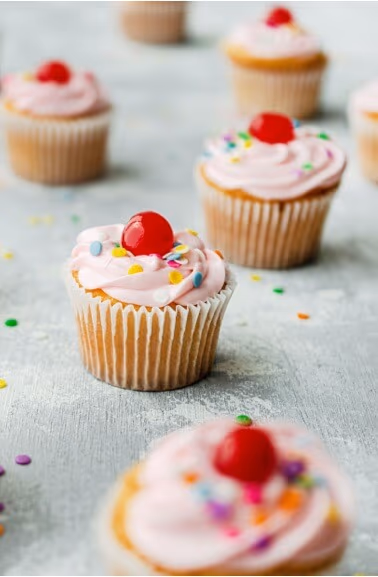 Pink frosted cupcakes decorated with red cherries and colorful sprinkles are arranged on a light grey surface.