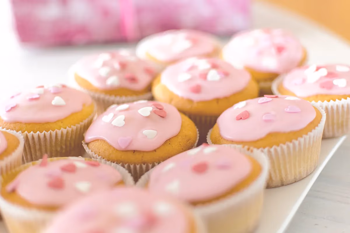 A collection of cupcakes topped with light pink frosting and small white and pink heart sprinkles, displayed on a white tray.