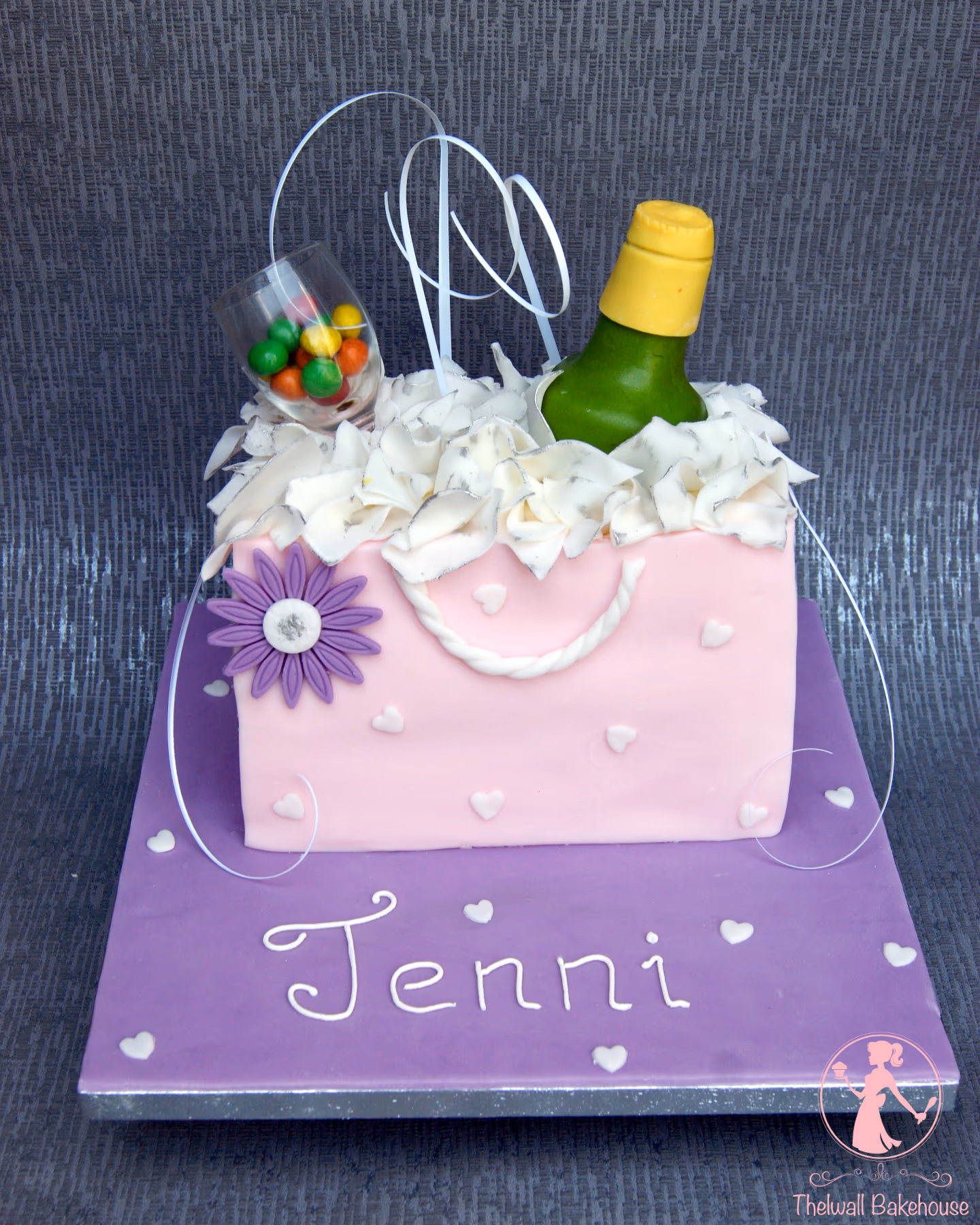 A personalized celebration cake for Jenni, shaped like a pink gift bag with white hearts, topped with a green champagne bottle, a glass filled with colorful candies, and white ruffled icing, all on a purple cake board.