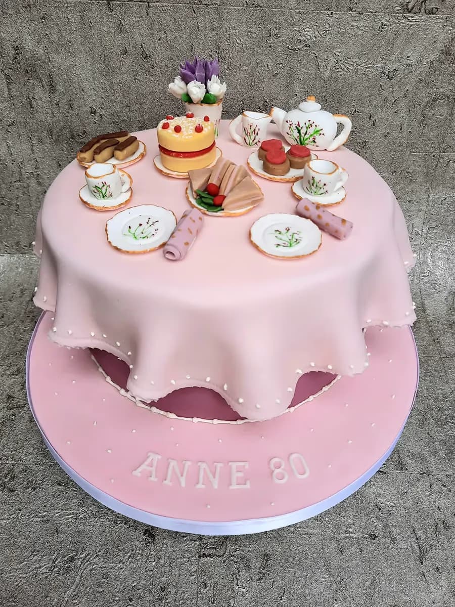 A two-tier pink birthday cake for Anne's 80th, designed as a miniature tea party with a draped tablecloth, tiny cakes, sandwiches, a teapot, cups, and a vase of flowers on top.