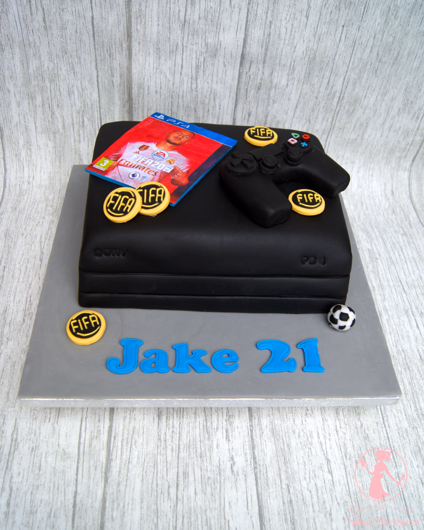 A black PlayStation console-shaped cake decorated with a FIFA 20 game cover, a black controller, yellow FIFA coins, a small soccer ball, and blue letters spelling 'Jake 21' on a grey cake board.