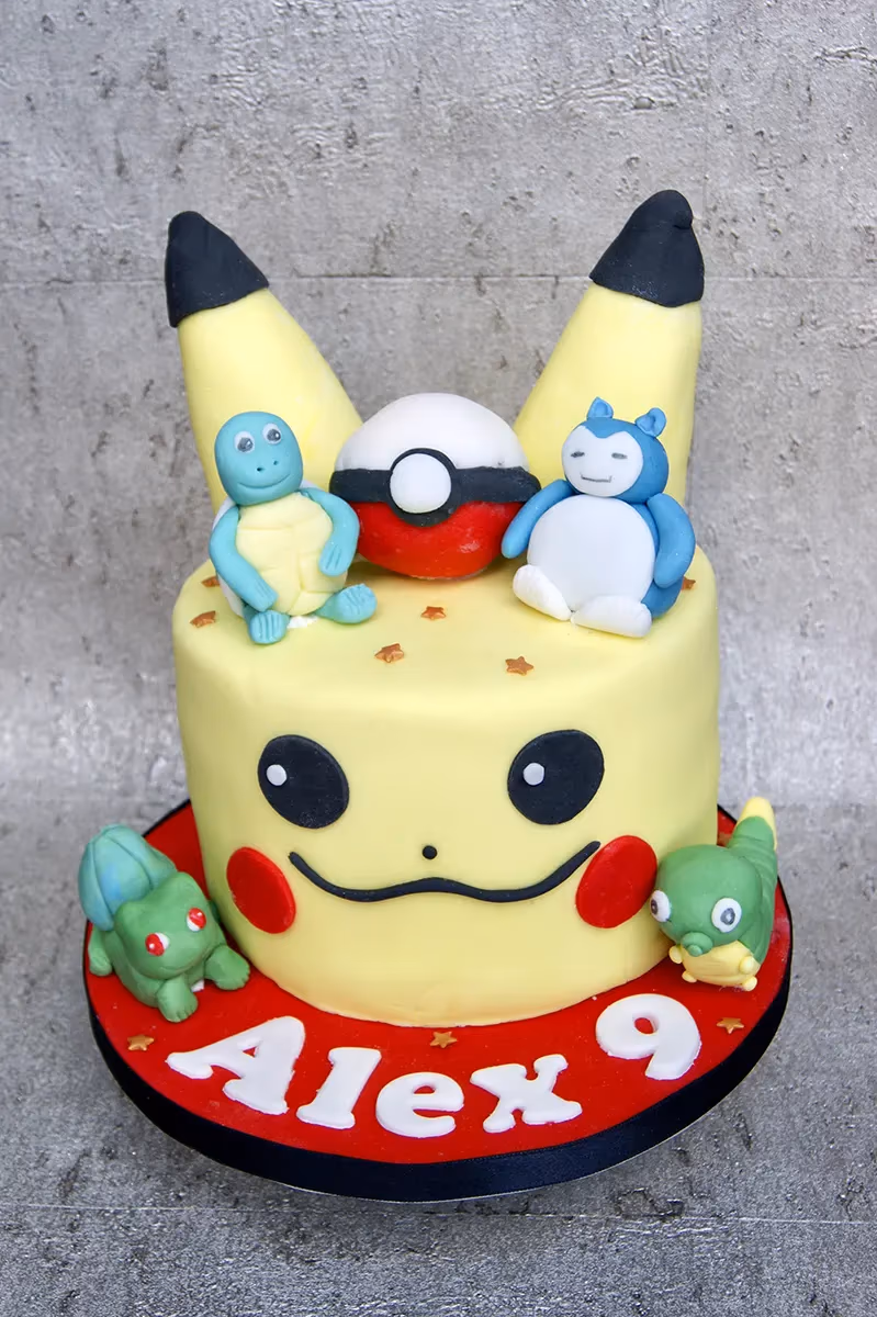 A vibrant yellow Pikachu-themed birthday cake featuring black ears, red cheeks, black eyes, and a smile. The top of the cake is decorated with fondant figures of Squirtle, a Poké Ball, and Snorlax, along with small golden star sprinkles. The red cake board at the bottom displays white letters spelling 'Alex 9', accompanied by fondant figures of Bulbasaur and a green worm-like Pokémon.