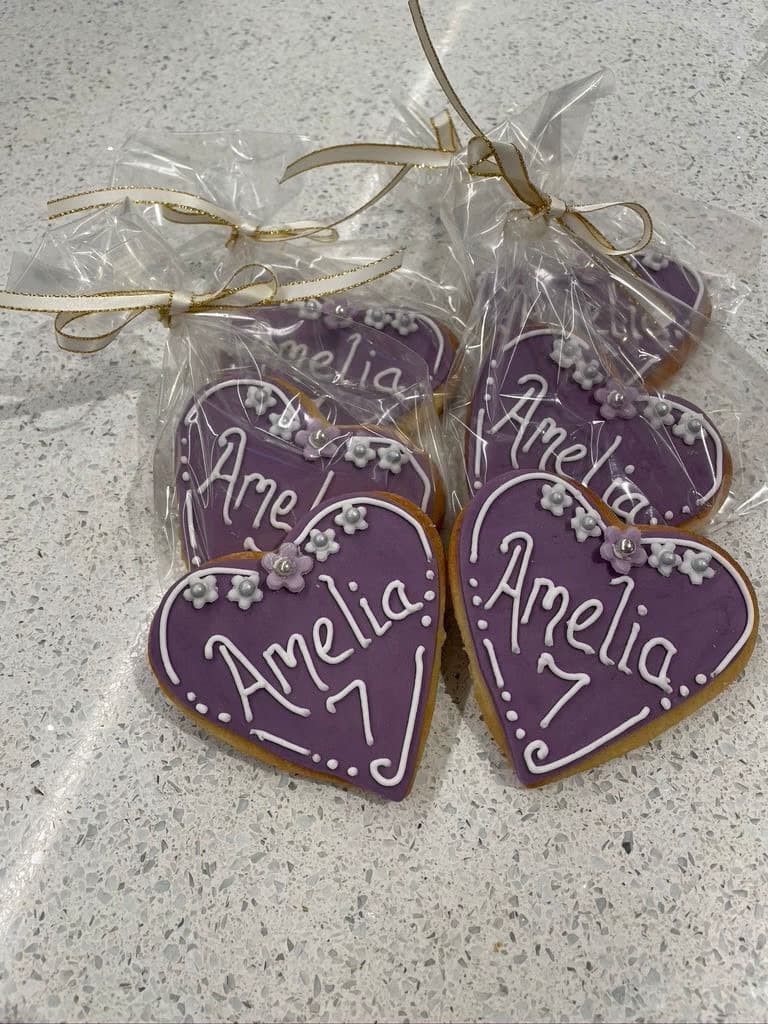Several heart-shaped biscuits with purple icing, featuring white piped details, white flowers, silver pearls, and the name 'Amelia' with the number '7', some individually packaged in clear bags tied with white and gold ribbons.