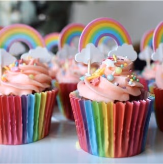 Close-up of several rainbow-themed cupcakes with pink frosting, rainbow sprinkles, white cloud toppers, and rainbow-striped paper liners.