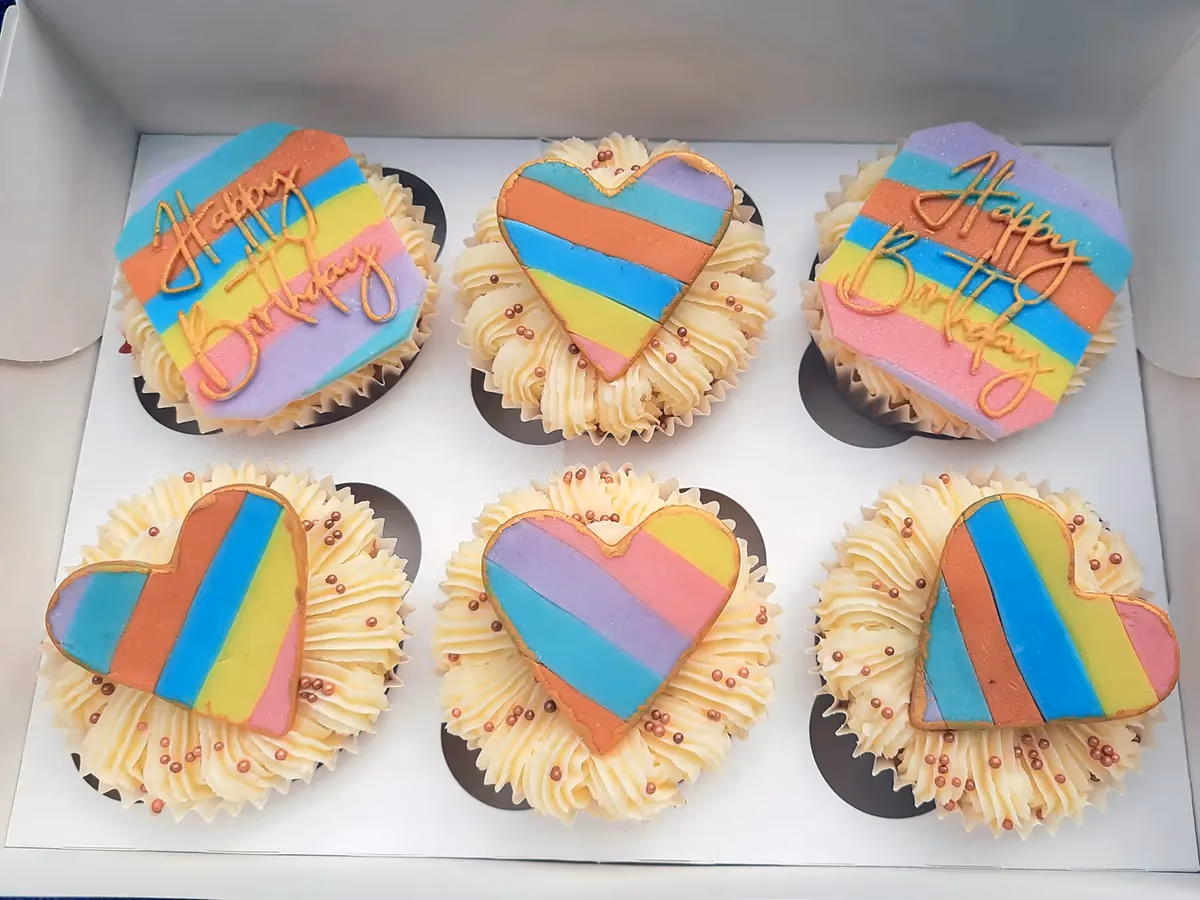 Six cupcakes in a white box, topped with light frosting and bronze sprinkles. Two cupcakes feature rainbow striped 'Happy Birthday' plaques, while four have rainbow striped heart-shaped plaques.