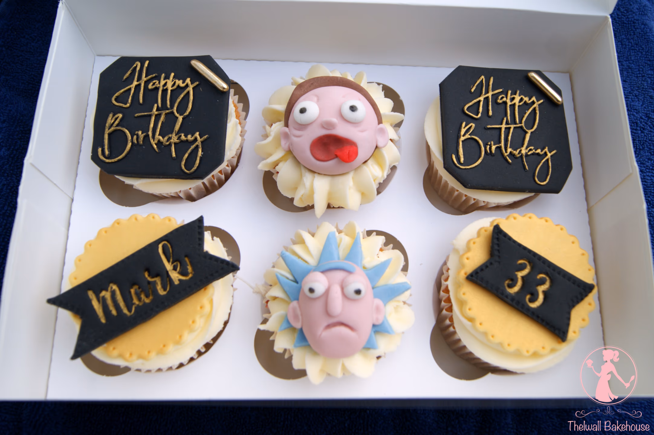 Six cupcakes in a white box, featuring two 'Happy Birthday' designs, an excited Morty face, a grumpy Rick face, a black banner with 'Mark' in gold, and a black banner with '33' in gold, all on cream frosting.