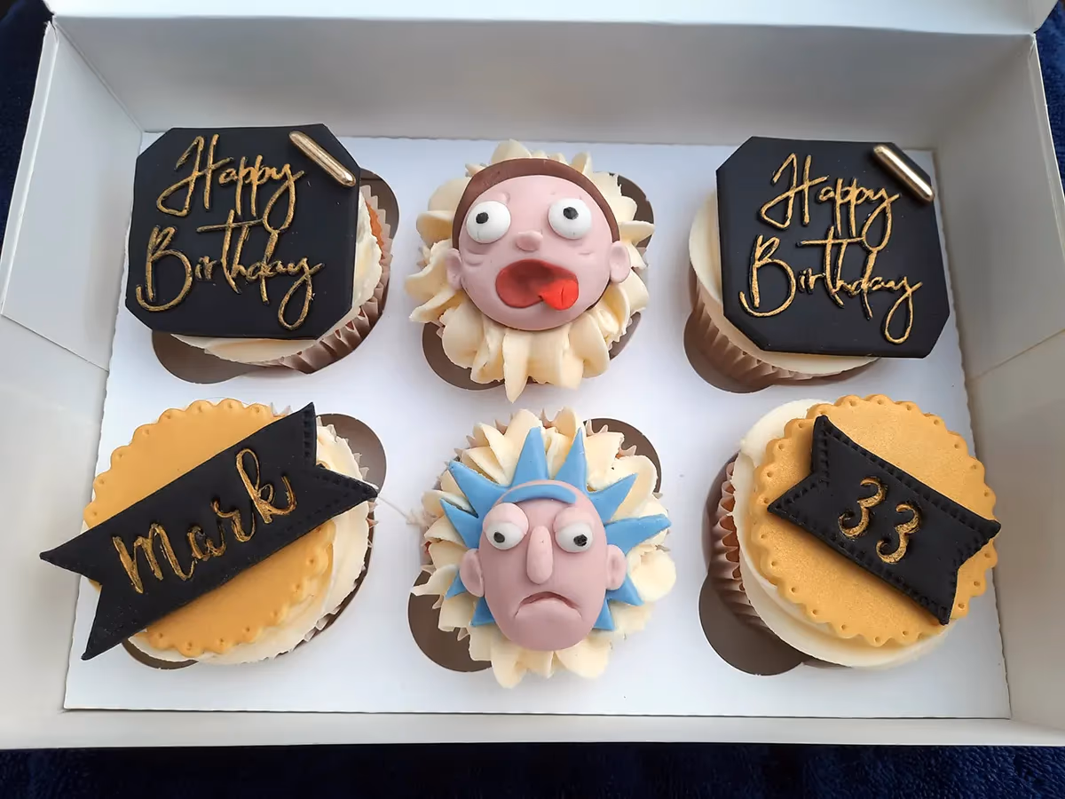 A white box containing six personalised birthday cupcakes. Two cupcakes feature black octagonal toppers with 'Happy Birthday' in gold script. Two cupcakes have edible fondant figures of Rick and Morty characters. The remaining two cupcakes have yellow scalloped bases, one with a black banner reading 'Mark' in gold, and the other with '33' in gold.