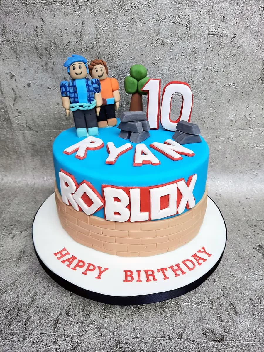 A round, two-tier Roblox-themed birthday cake with a blue top tier and a light brown brick-patterned bottom tier, decorated with two Roblox character figurines, the number 10, grey block stacks, and the words 'RYAN' and 'ROBLOX'. The white cake board reads 'HAPPY BIRTHDAY' in red letters.