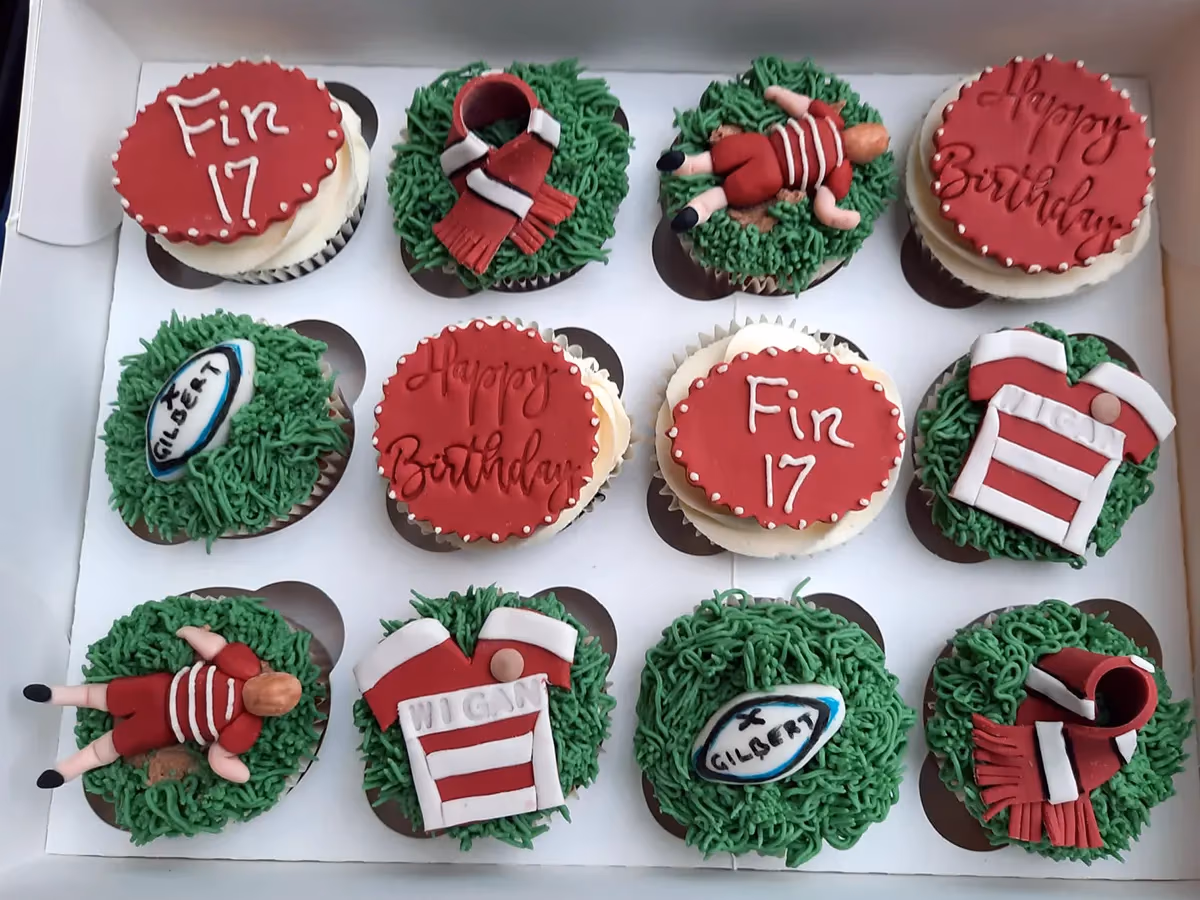 A dozen rugby-themed birthday cupcakes in a white box, featuring green grass frosting, red and white decorations including rugby jerseys, player figures, rugby balls, scarves, and messages 'Fin 17' and 'Happy Birthday'.