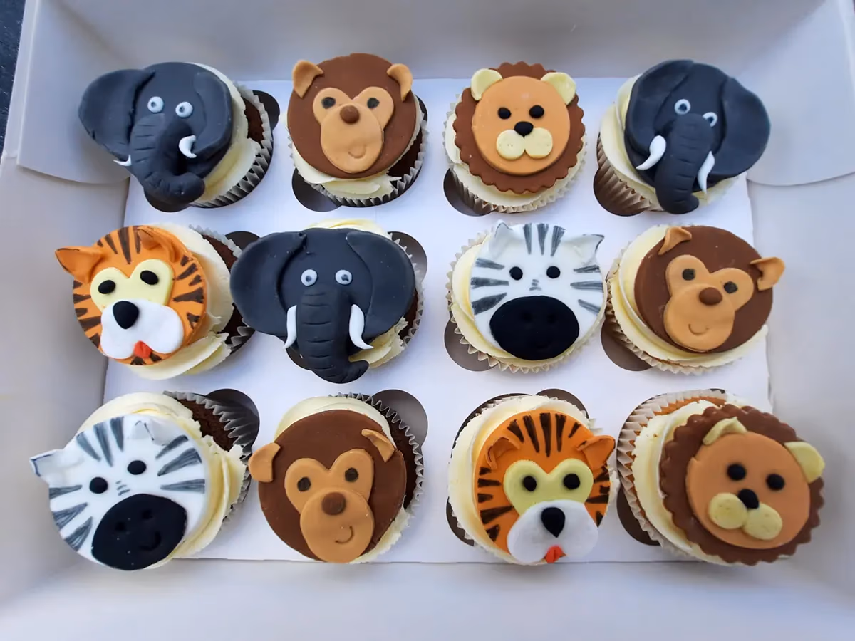 A white box filled with twelve cupcakes, each decorated with a different safari animal face made from fondant. The assortment includes elephants, monkeys, lions, tigers, and zebras.
