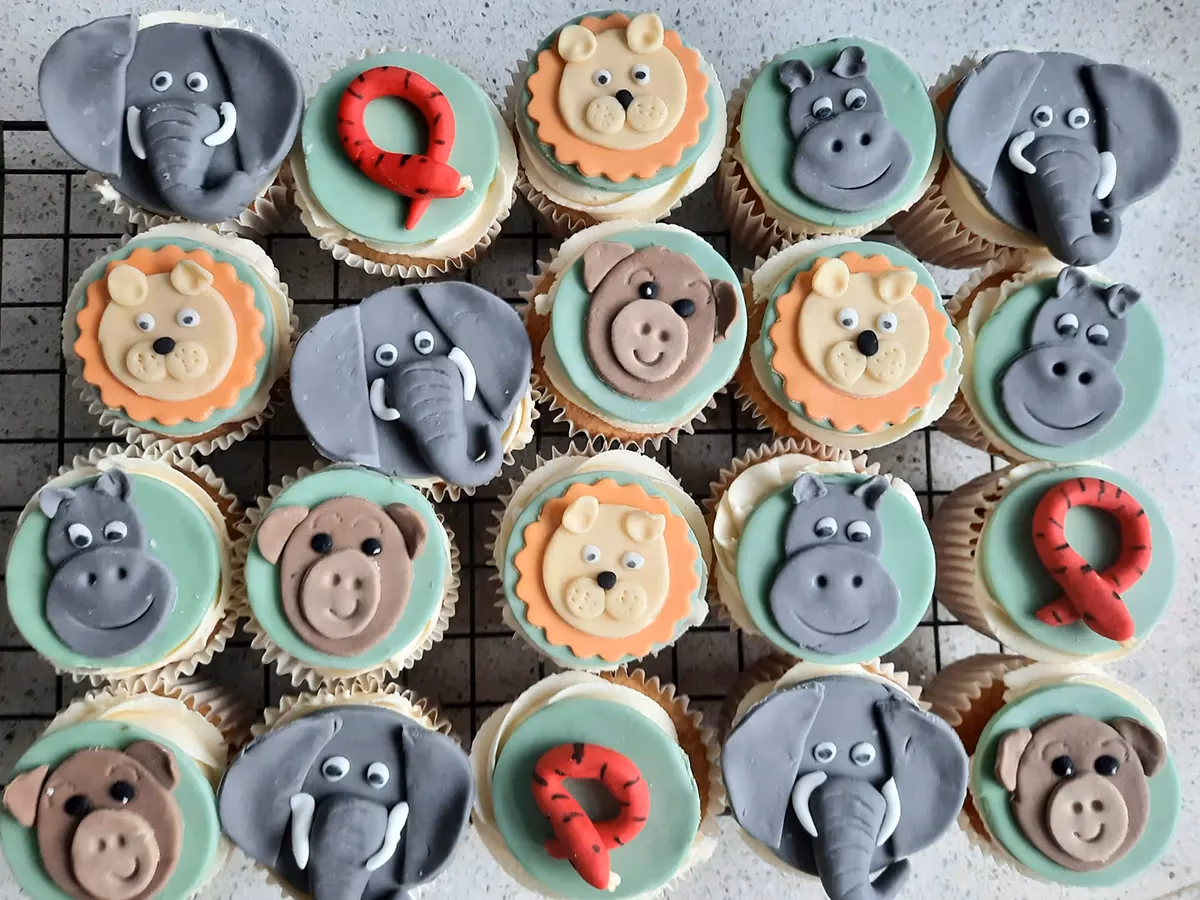 An overhead view of numerous cupcakes decorated with fondant toppers of various safari animals, including elephants, lions, hippos, monkeys, and snakes, arranged on a black cooling rack.