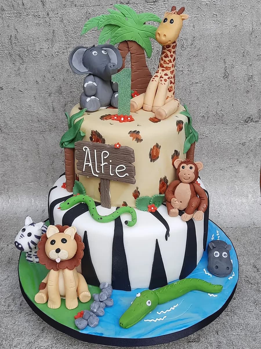 A two-tier safari-themed first birthday cake decorated with fondant animal figures including an elephant, giraffe, monkey, lion, zebra, hippo, crocodile, and snake. The top tier features a leopard print, a palm tree, and the number '1', while the bottom tier has a zebra stripe pattern with a water feature and a sign reading 'Alfie'.