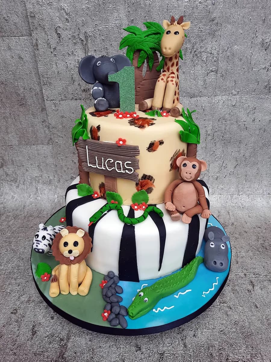 A vibrant two-tier safari-themed birthday cake for a first birthday, featuring numerous edible animal figurines. The top tier includes an elephant, a giraffe, a monkey, a green glittery number '1', and a 'Lucas' sign, surrounded by jungle foliage. The bottom tier has black and white zebra stripes, and the cake board is decorated with a zebra, a lion, a hippo, and a crocodile in a blue water pond.