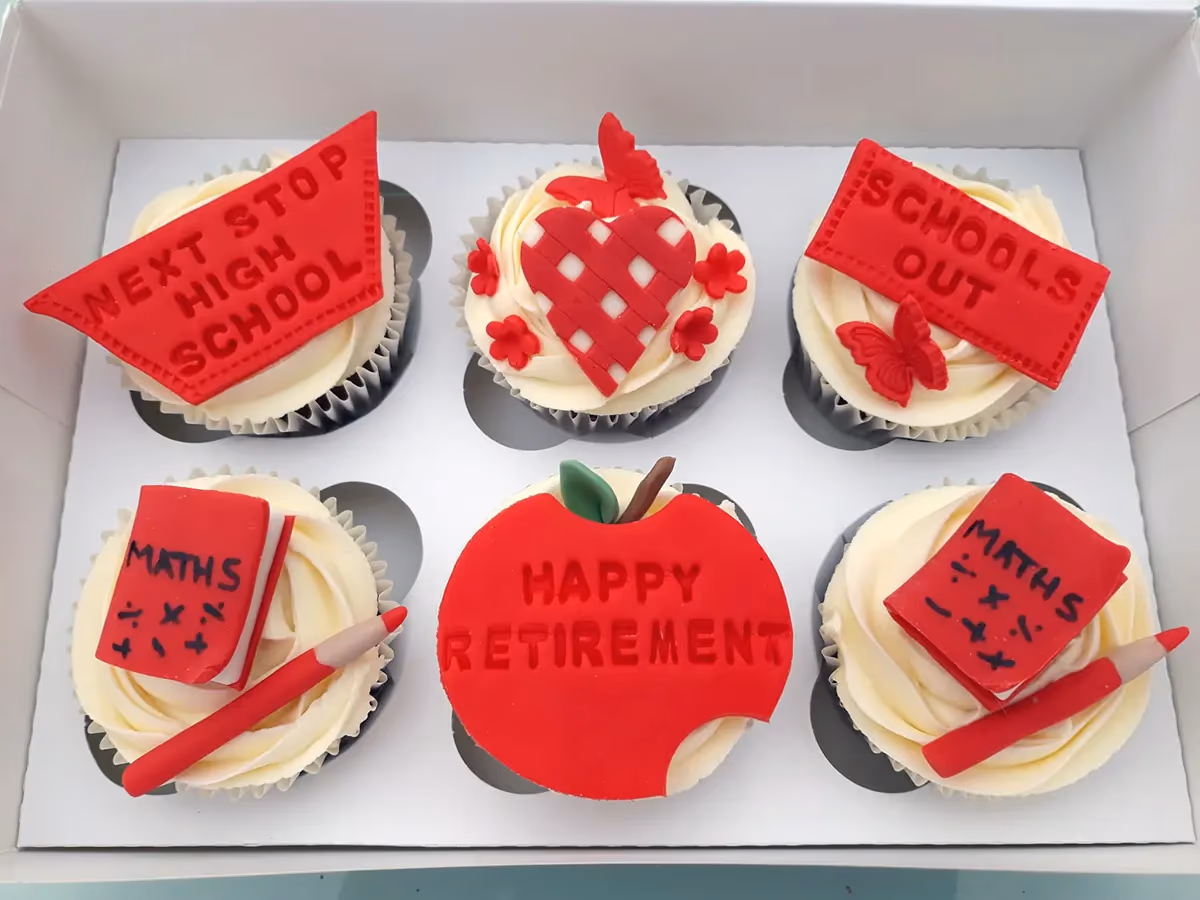 Six school-themed cupcakes in a white box, decorated with red fondant toppers. These include an apple with 'HAPPY RETIREMENT', a flag reading 'NEXT STOP HIGH SCHOOL', a rectangle with 'SCHOOLS OUT', a heart, and two cupcakes with 'MATHS' books and pencils.