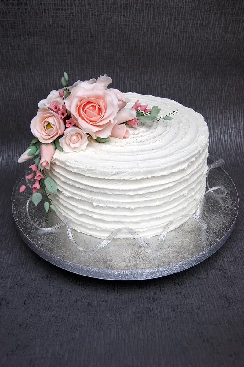 A single-tier white wedding cake with ridged buttercream frosting, adorned with a cluster of pink sugar roses and green foliage, on a silver cake board with a silver ribbon.