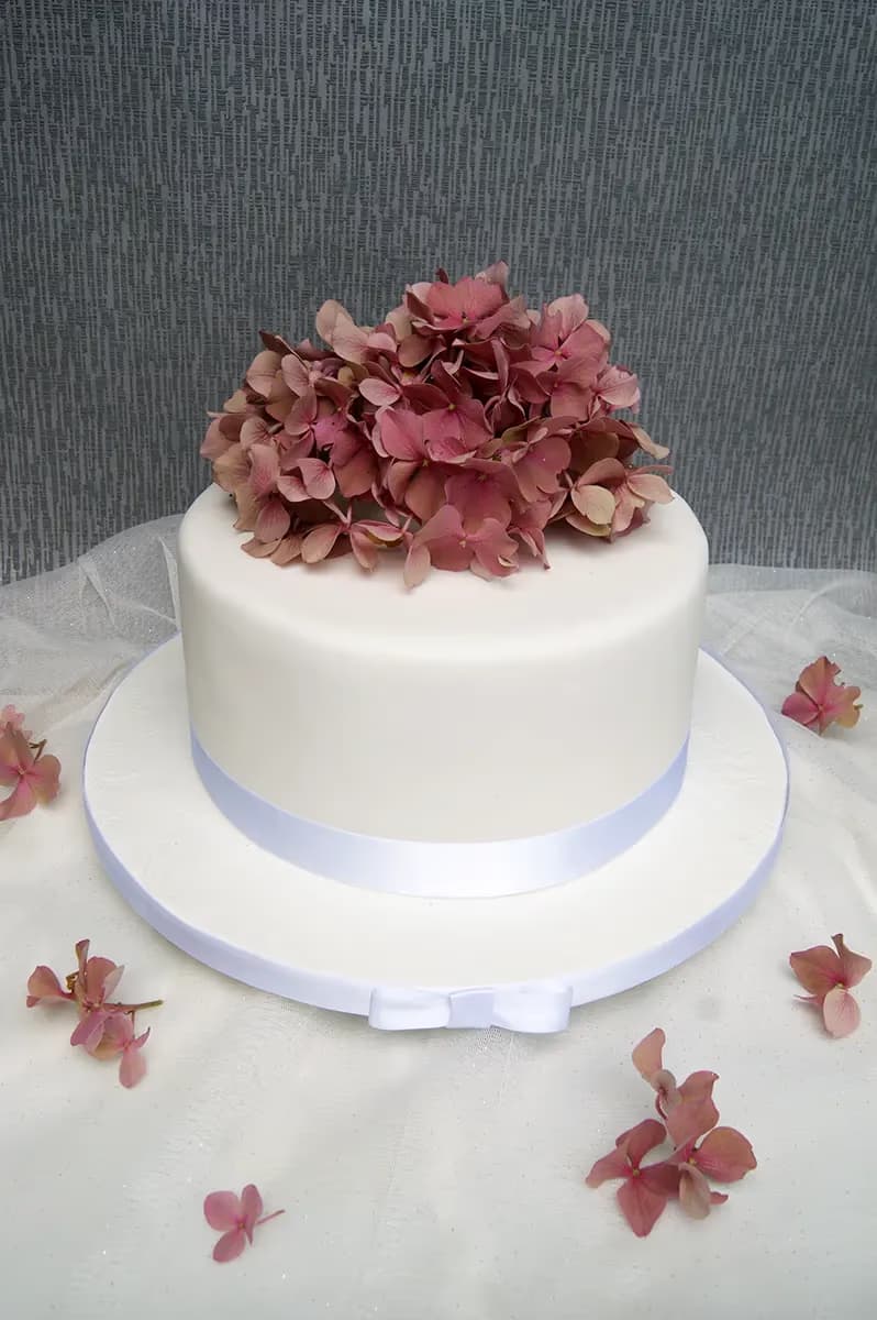 A single-tier white cake decorated with a large cluster of pink hydrangea flowers on top, sitting on a white cake board with a white ribbon and bow, surrounded by scattered pink petals on a white sheer tablecloth.