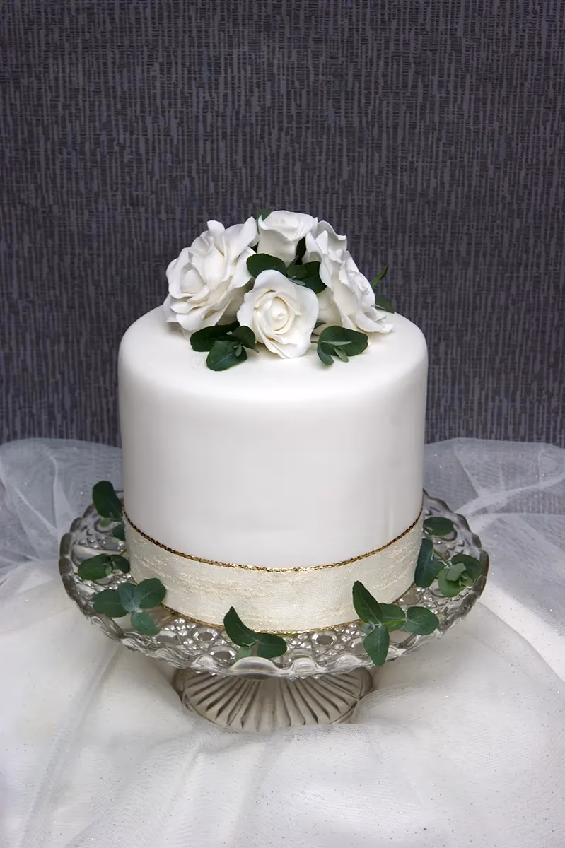 A single-tier white wedding cake, decorated with white sugar roses and eucalyptus leaves on top, and a cream ribbon with gold trim around its base, sitting on a clear glass cake stand.
