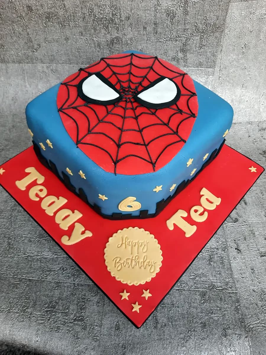 A square birthday cake featuring a blue base and a red Spiderman face on top with black web lines and white eyes. The cake is decorated with gold stars and a gold number 6, and sits on a red cake board with 'Teddy Ted' written in gold letters and a gold 'Happy Birthday' plaque.