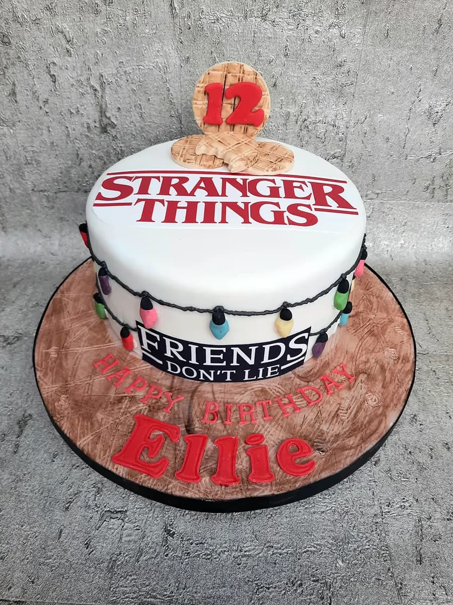 A white round Stranger Things themed birthday cake for Ellie's 12th birthday, featuring the show's logo, a string of colorful lights, and waffle decorations, with 'Happy Birthday Ellie' written in red on a wood-textured cake board.