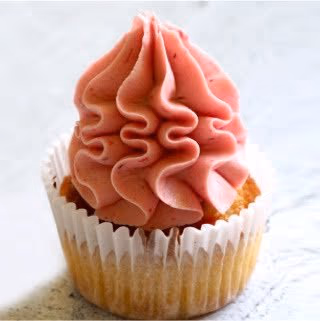 A single cupcake in a white paper liner, topped with a tall, elegant swirl of bright pink frosting, speckled with small pieces of fruit.