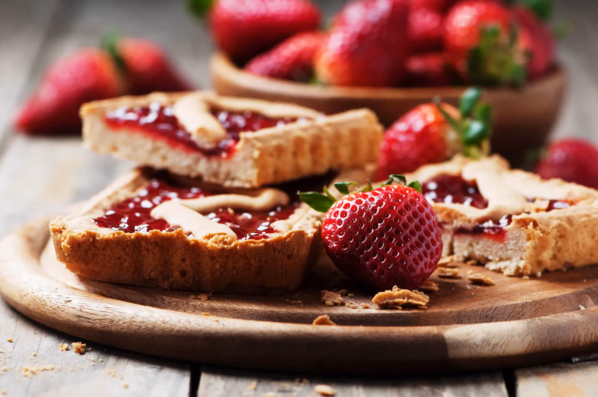 Homemade strawberry jam tarts with pastry strips on top, served on a rustic wooden board alongside fresh whole strawberries.