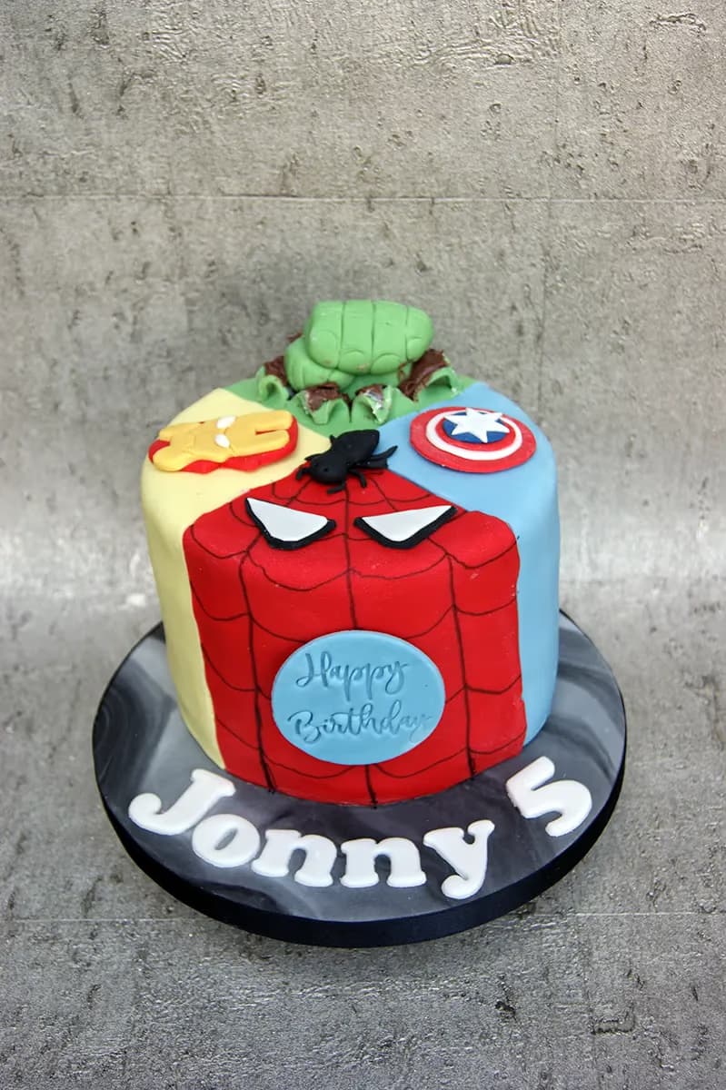 A round birthday cake decorated with four superhero themes: Hulk, Iron Man, Spider-Man, and Captain America. The cake has a light blue circle on the Spider-Man section with 'Happy Birthday' written on it, and the name 'Jonny' and the number '5' are spelled out in white letters on the black and grey marbled cake board.