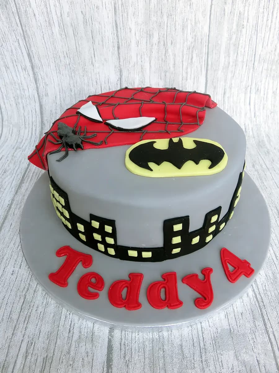 A single-tier grey fondant birthday cake featuring a red Spiderman mask, a black and yellow Batman logo, a black city skyline around the base, and the name 'Teddy 4' in red letters on the cake board.