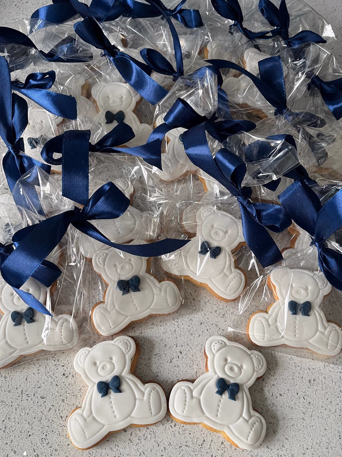A close-up view of numerous white teddy bear cookies, each featuring a small blue bow tie. Most are individually wrapped in clear plastic bags tied with dark blue satin ribbons, while a few unwrapped cookies are visible in the foreground on a light grey speckled surface.