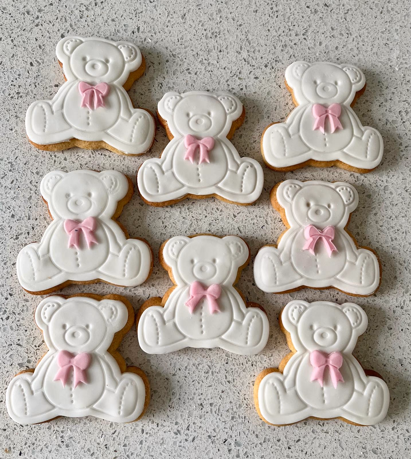 Nine white teddy bear shaped sugar cookies, each decorated with a small pink bow, laid out on a speckled light grey countertop.
