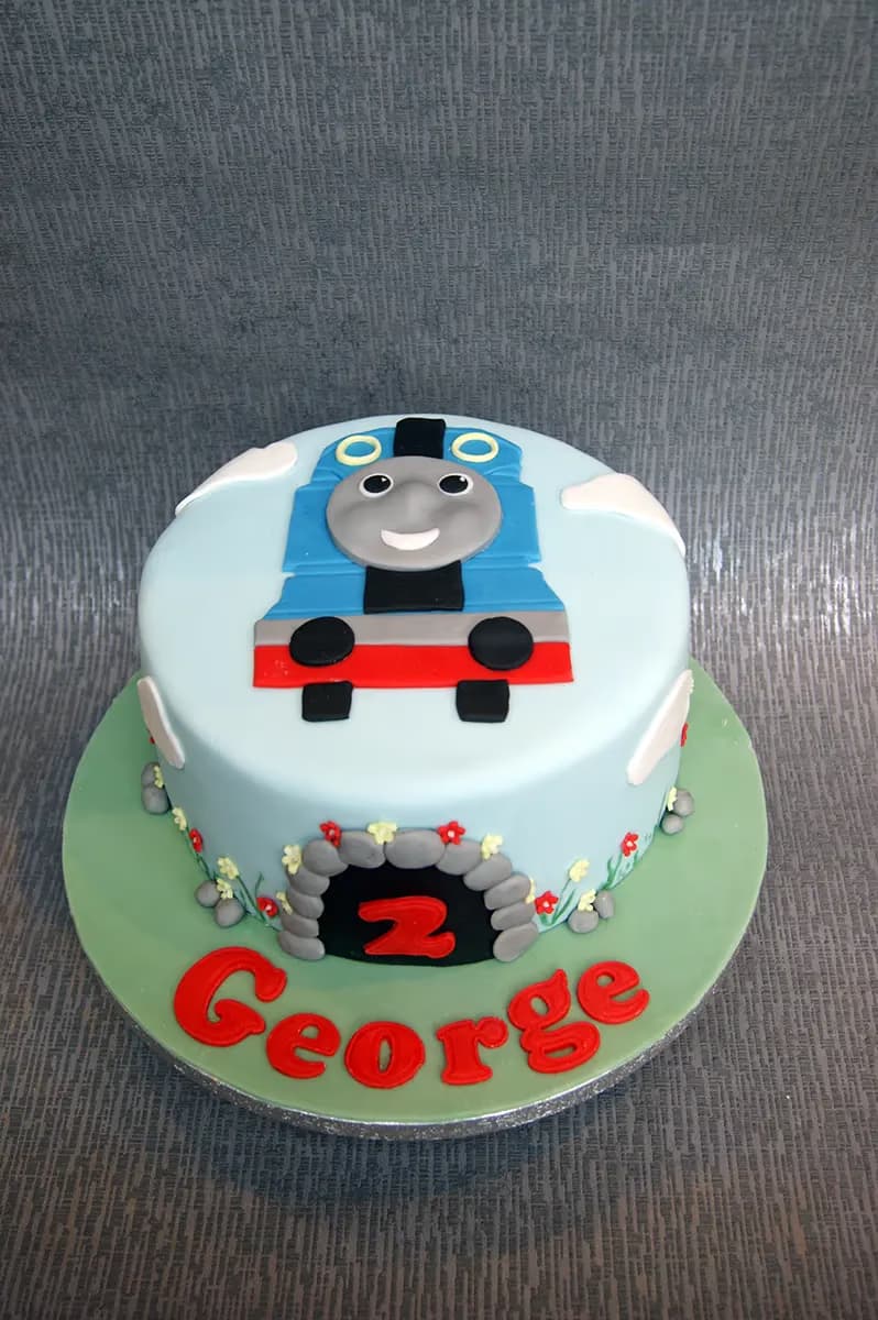 A light blue round birthday cake decorated with Thomas the Tank Engine on top, a grey tunnel with the number '2' on the side, and the name 'George' in red letters on the green cake board.