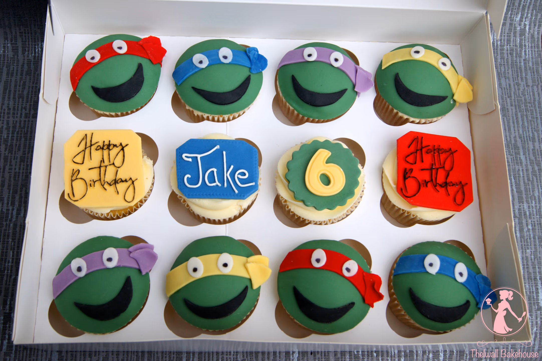A white box containing twelve cupcakes, featuring eight green Teenage Mutant Ninja Turtles themed cupcakes with red, blue, purple, and yellow masks, and four birthday cupcakes with 'Happy Birthday' text, the name 'Jake', and the number '6'.