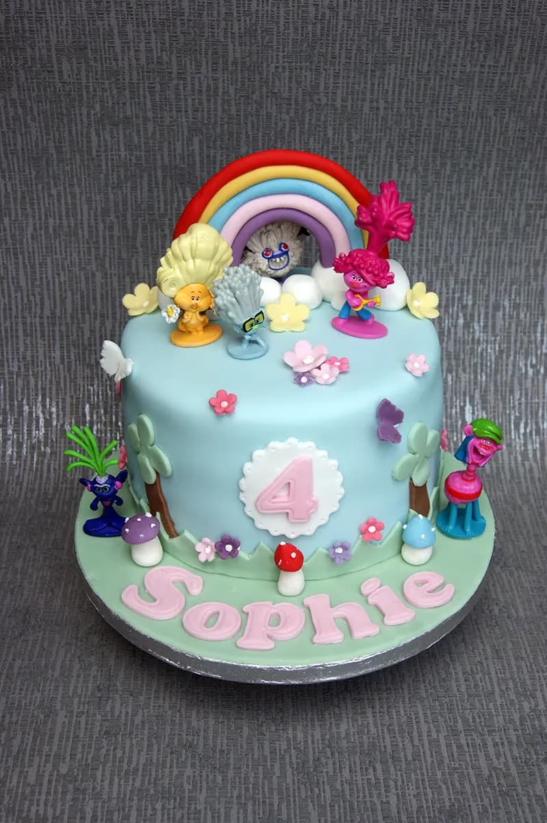 A vibrant single-tier Trolls-themed birthday cake for Sophie's 4th birthday, featuring a light blue fondant base, a colorful rainbow, and various Trolls characters like Poppy, Branch, and Guy Diamond, along with edible flowers, mushrooms, and palm trees. The name Sophie is spelled out in pink letters on the base, and the number 4 is on the side of the cake.