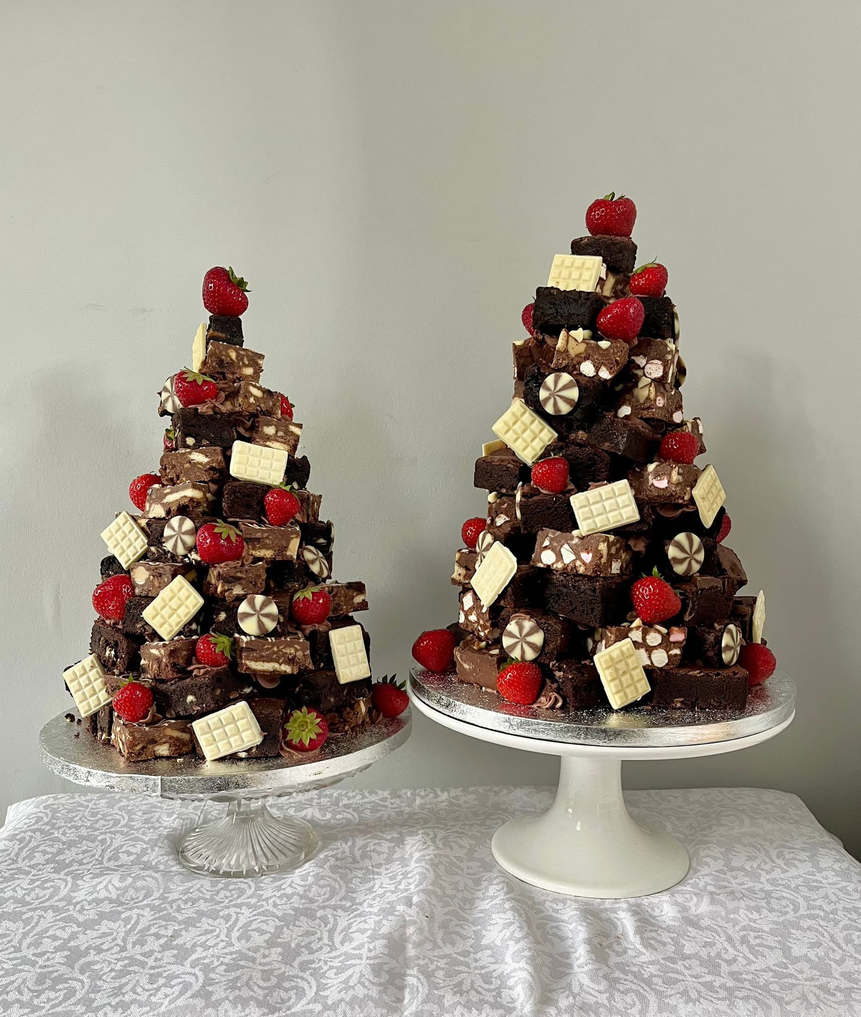 Two festive brownie stacks, resembling trees, decorated with fresh strawberries, white chocolate squares, and chocolate swirl candies, presented on cake stands.