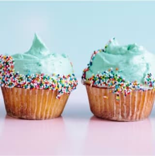 Two vanilla cupcakes topped with light blue frosting and colorful sprinkles, set against a pastel pink and blue background.