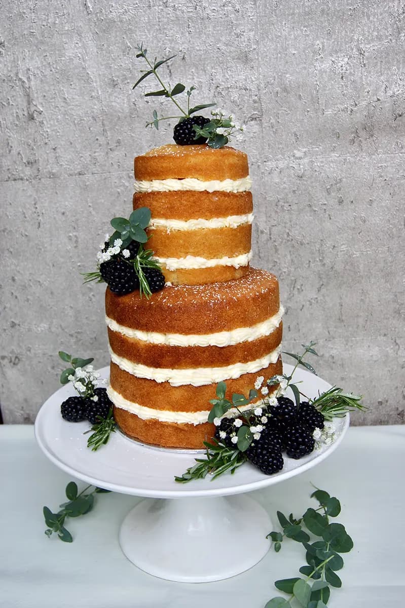 A two-tier naked cake with visible golden cake layers and white cream filling, adorned with fresh blackberries, green eucalyptus, rosemary, and small white flowers, presented on a white cake stand.