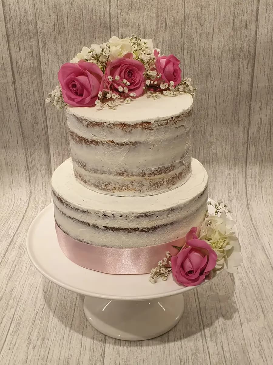A two-tier naked cake featuring visible cake layers and thin white frosting, decorated with pink roses, white flowers, baby's breath, and a pink satin ribbon around the base.