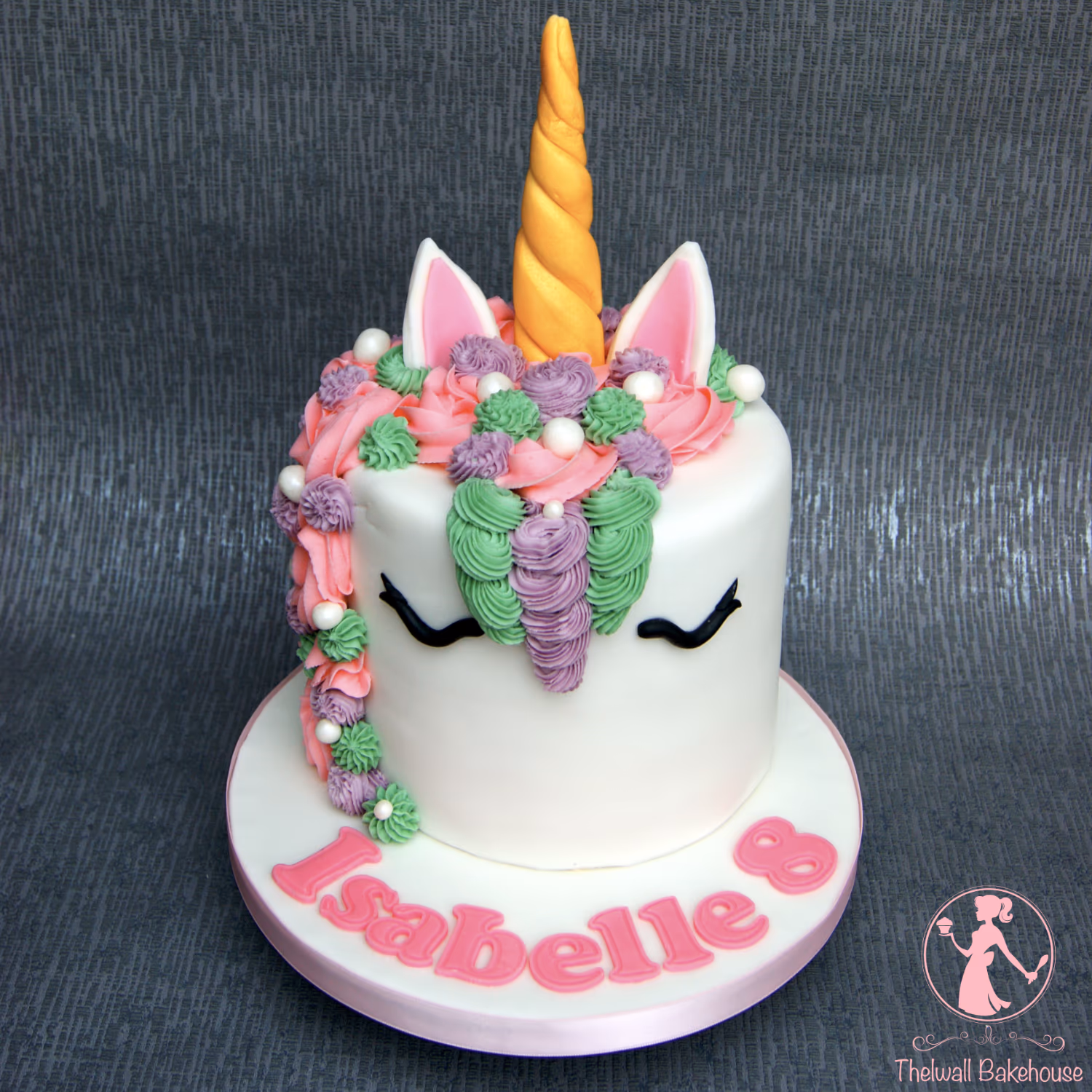 A single-tier white fondant unicorn cake with a gold horn, pink and white ears, and a mane of pink, purple, and green piped frosting with white pearls. The cake board features pink fondant letters spelling 'Isabelle 8'.