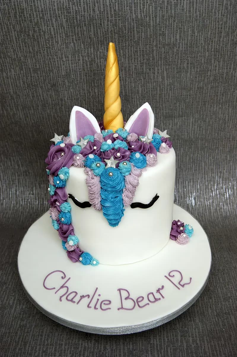 White unicorn cake decorated with a gold horn, purple and blue piped buttercream mane with silver star sprinkles, and 'Charlie Bear 12' written in purple on the white cake board.