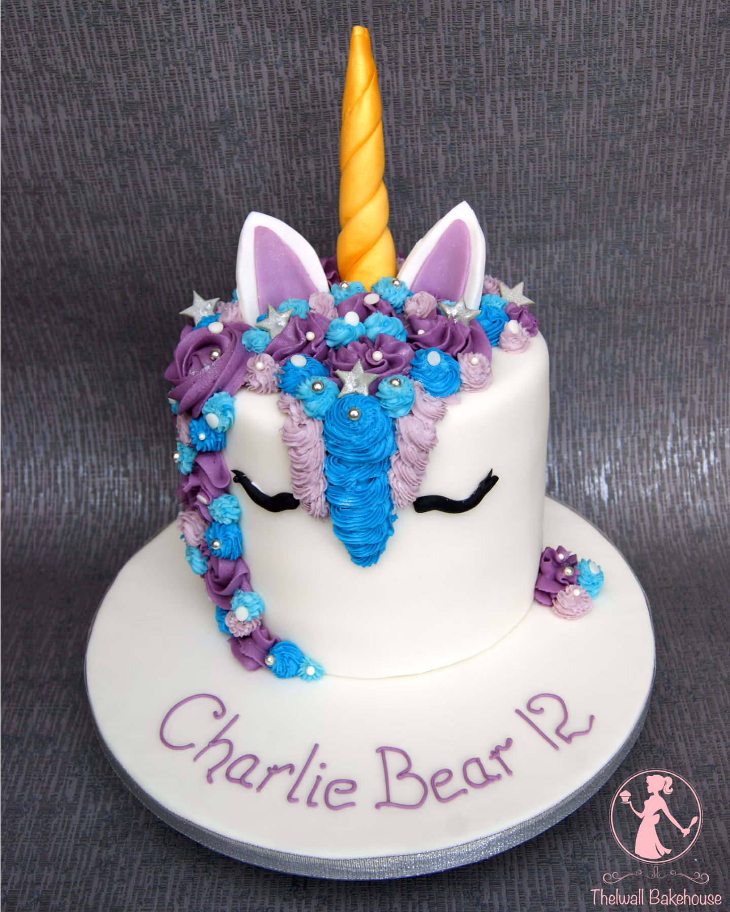 White fondant unicorn cake with a gold horn, purple and blue icing mane and flowers, silver stars, and silver beads, with 'Charlie Bear 12' written on the board.