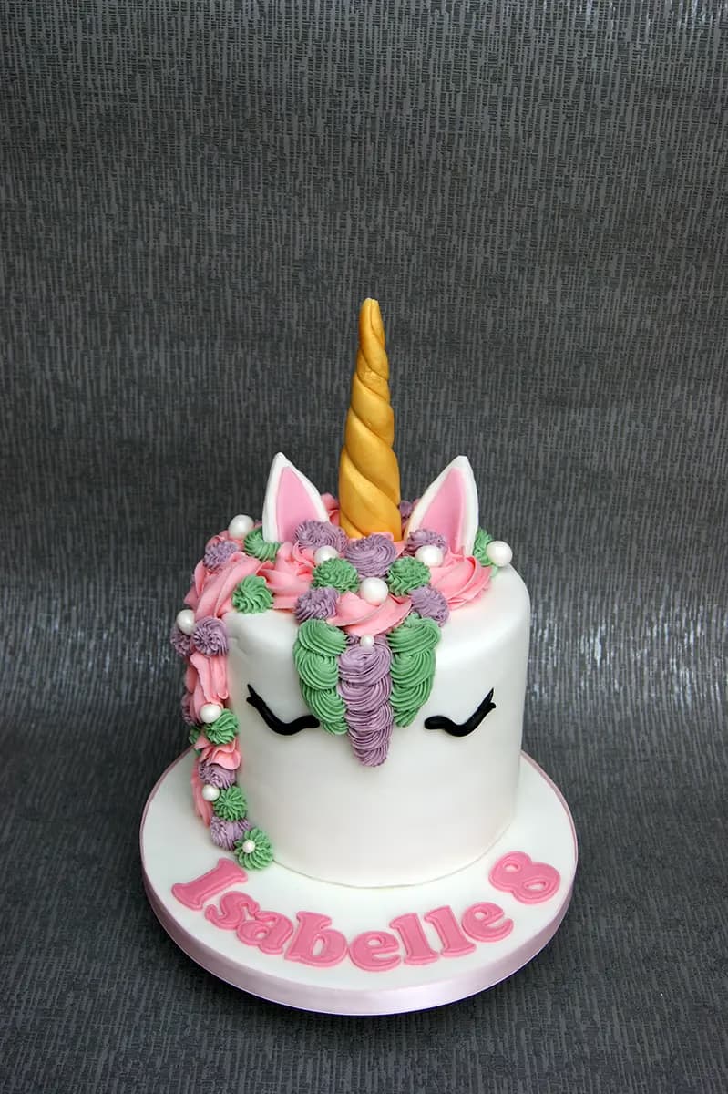 A white unicorn cake with a golden horn, pink and white ears, and a colorful piped mane in shades of pink, green, and purple, adorned with small white pearls. The cake board reads 'Isabelle 8' in pink letters.