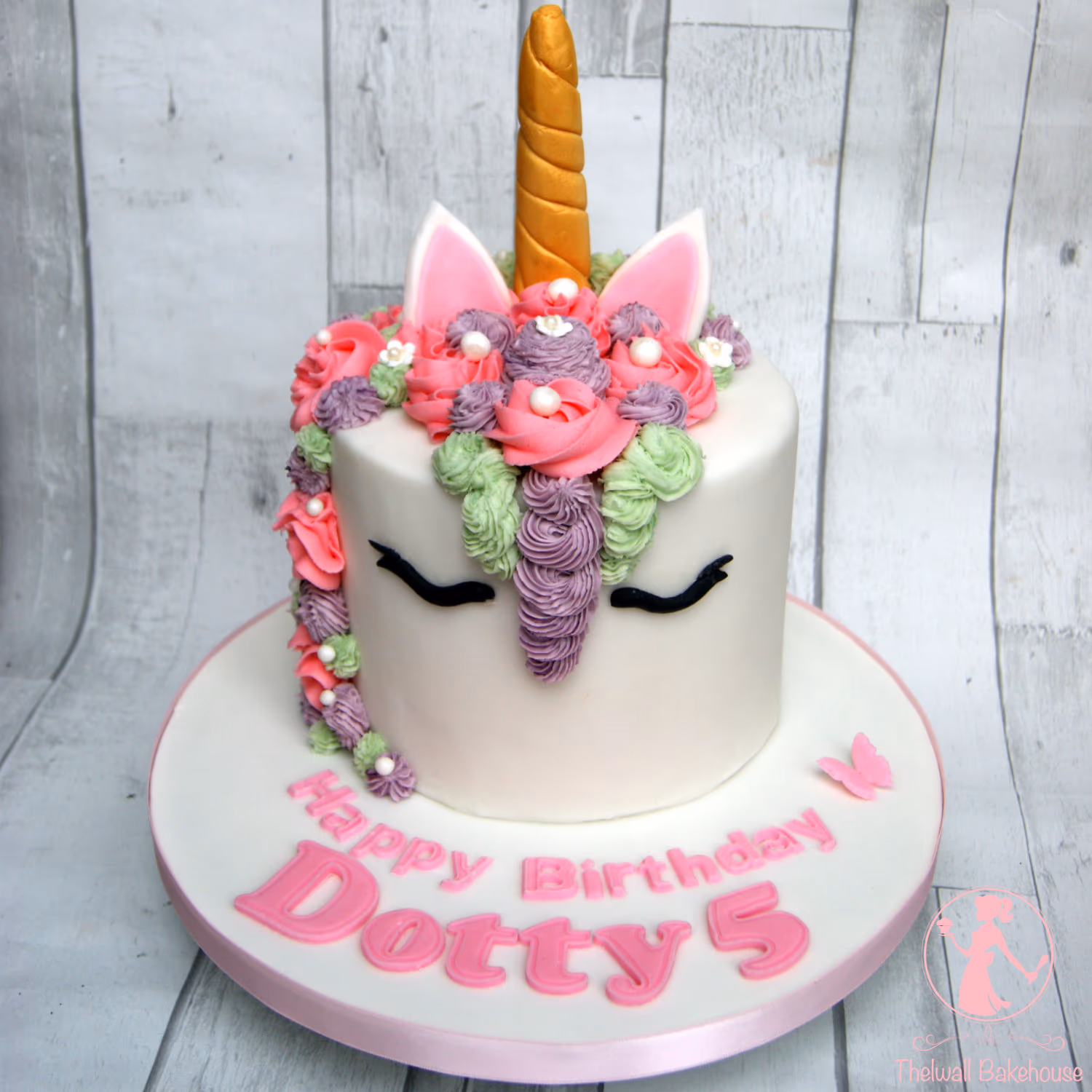 A white single-tier unicorn cake with a golden horn, pink and white ears, and a vibrant piped mane in pink, purple, and green, decorated with white pearls. Black piped eyes are on the front. The cake board displays 'Happy Birthday Dotty 5' in pink letters and a small pink butterfly.