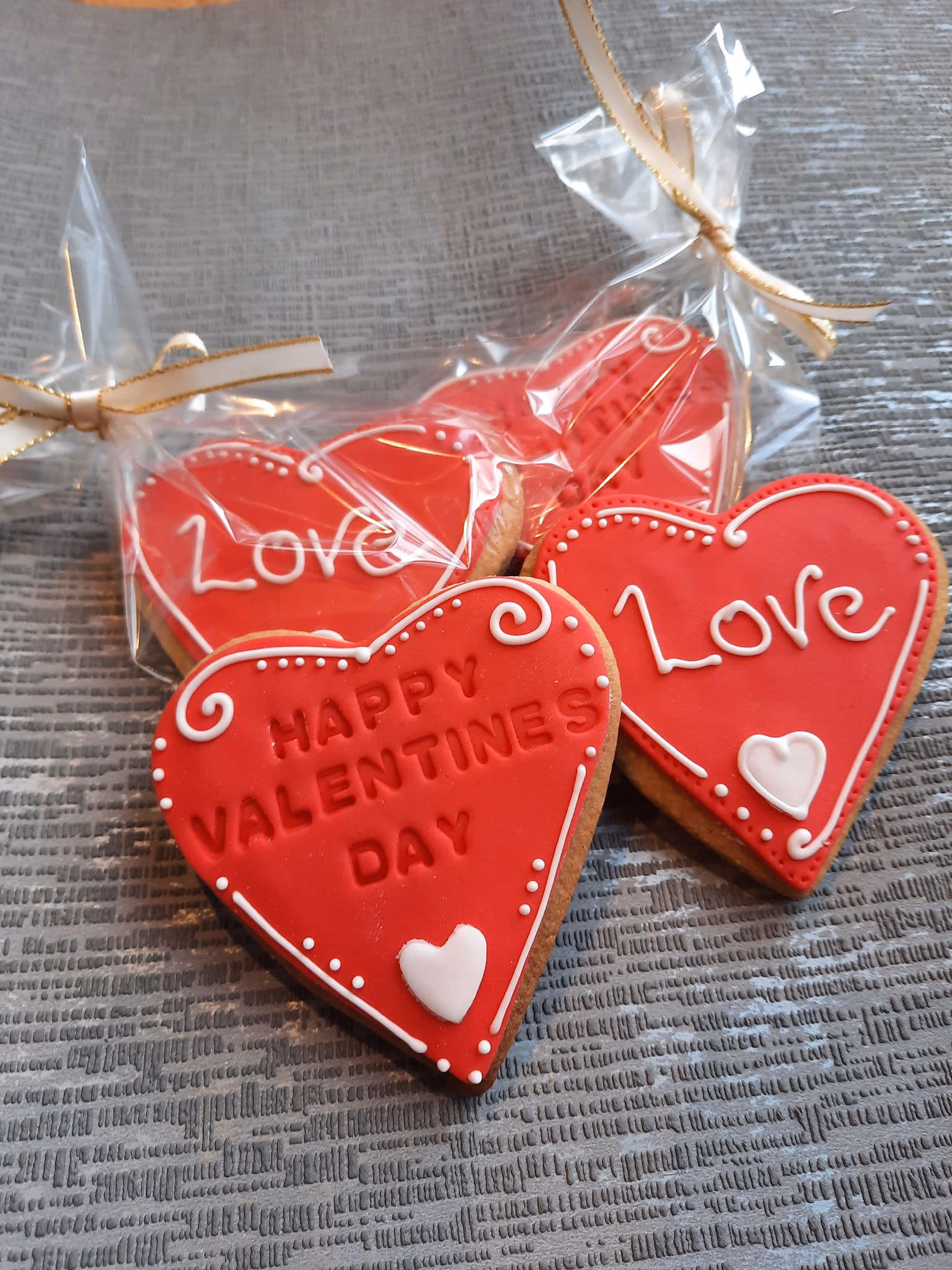 Several red heart-shaped Valentine's Day cookies decorated with white icing, some featuring 'Love' and one 'Happy Valentines Day', with some packaged in clear bags tied with gold ribbons.