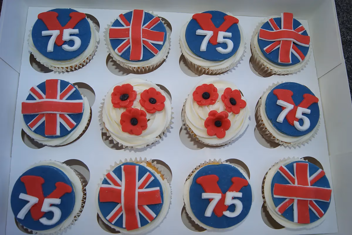 A dozen cupcakes in a white box, featuring three different designs: some with a blue fondant top displaying a red 'V' and white '75' for VE Day 75, others with a red and white Union Jack flag, and some with white frosting topped with three red poppy flowers.